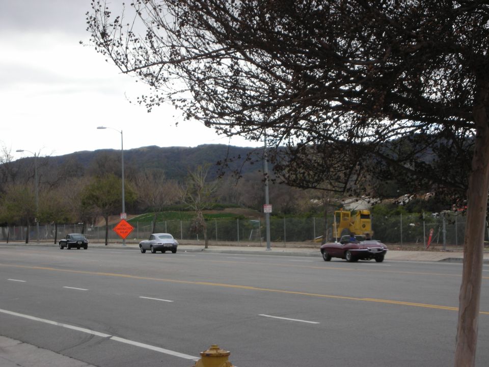 Dark red convertible and cars driving on a road below hills and bare trees
