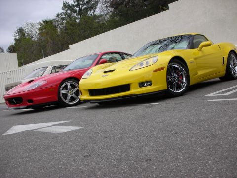 1998 Ford Crown Victoria - yellow Chevrolet Corvette Z06 and a red Ferrari coupe sit side by side in a lot against a plain white wall.