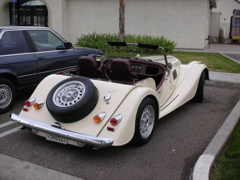 1998 Ford Crown Victoria - cream Morgan roadster with red interior shows its rear-mounted spare wheel and chrome bumper while parked at a curb.