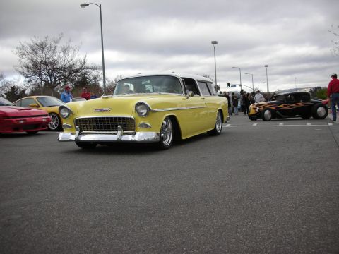 1998 Ford Crown Victoria - lowered yellow mid-1950s Chevrolet Nomad two-door wagon with chrome bumper and polished wheels rolls across a parking-lot car show.