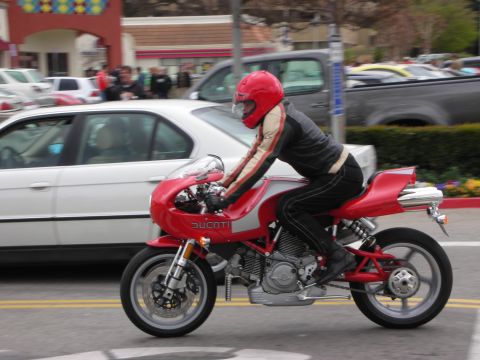 1998 Ford Crown Victoria - leather-clad rider in a red helmet rides a red Ducati sportbike along a street past parked cars.