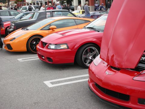 1998 Ford Crown Victoria - lineup of parked sports cars led by an orange Lamborghini, a red Mustang Cobra and a red Corvette with its hood up.
