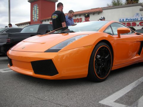 1998 Ford Crown Victoria - orange Lamborghini Gallardo with black wheels and angular nose parked at a strip-mall car show with spectators behind.