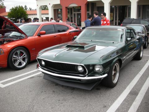 1998 Ford Crown Victoria - dark green late-1960s Ford Mustang fastback with chrome bumpers and a hood scoop parked at a strip-mall car show.