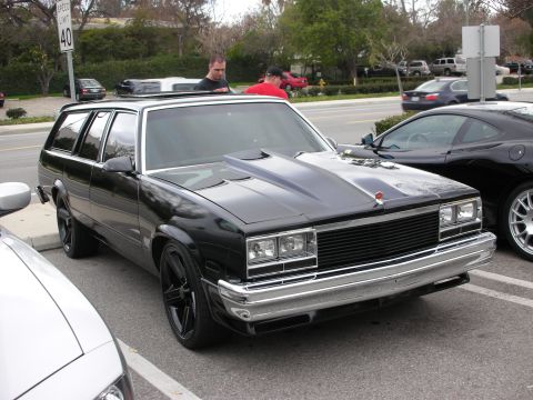 1998 Ford Crown Victoria - lowered black 1980s GM full-size station wagon with cowl hood and black wheels parked at a tree-lined lot.
