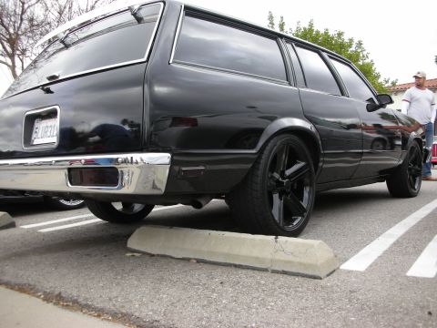 1998 Ford Crown Victoria - Low rear-quarter shot of a lowered black station wagon on black wheels straddling a parking curb, California plate visible.