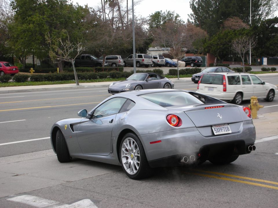 Rear three-quarter of silver Ferrari 599 coupe on a tree-lined street with traffic