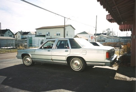 1989 Ford LTD Crown Victoria - A silver full-size Ford sedan shown in profile from the driver's side, parked on pavement with houses, a chain-link fence, and a red-and-bl…