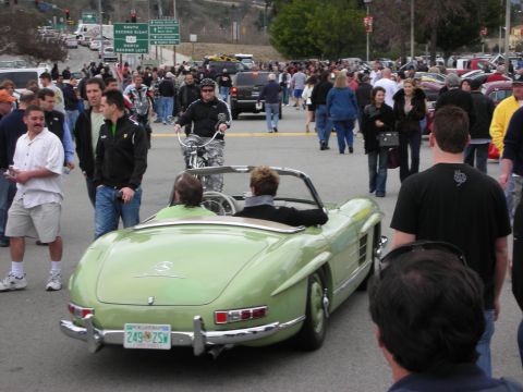 1998 Ford Crown Victoria - green classic Mercedes 300SL roadster carries two people through a dense crowd of spectators at an outdoor car event.