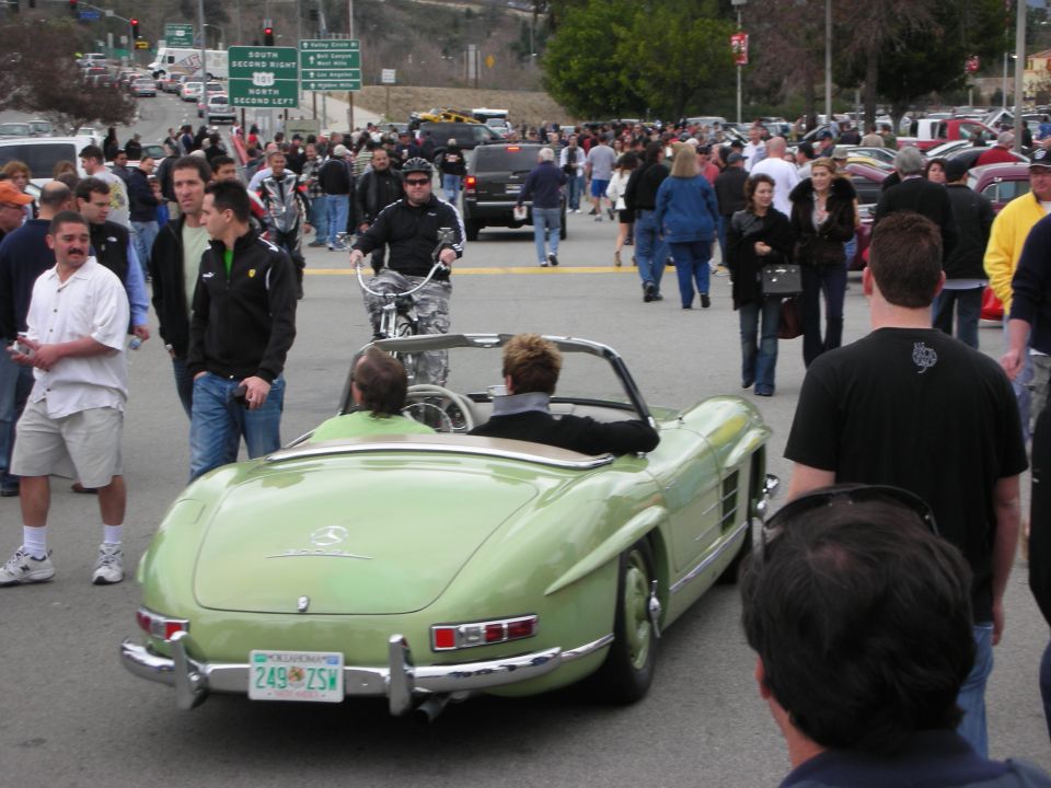 Rear of green Mercedes 300SL roadster with two occupants in a crowd at a car show