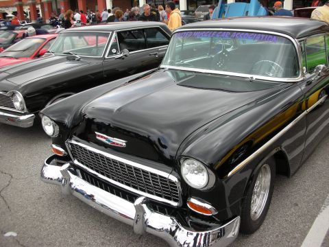 1998 Ford Crown Victoria - black 1955 Chevrolet with a chrome grille and bumper sits beside a red car in a crowded outdoor car show.