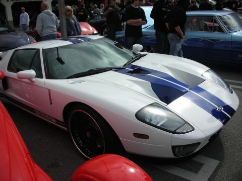 1998 Ford Crown Victoria - white Ford GT with blue dual stripes and chrome wheels sits among red and other cars at a crowded car show.