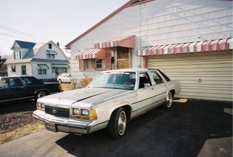 1989 Ford LTD Crown Victoria - A white/silver Ford full-size sedan photographed from the front three-quarter angle, parked on a driveway beside a house with a striped awn…