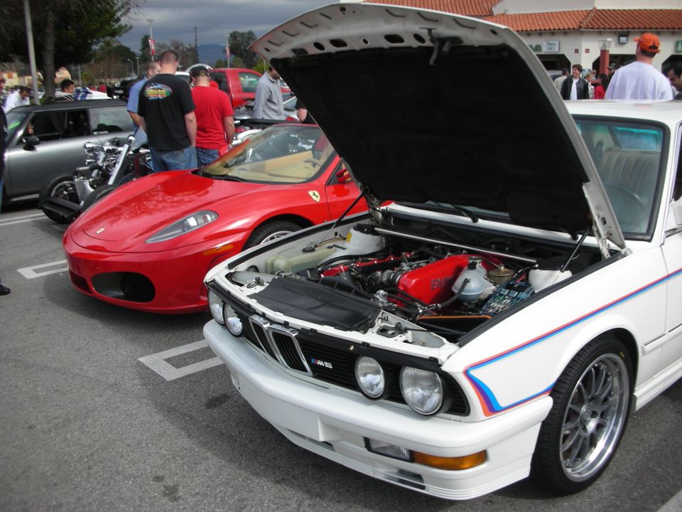 White BMW M-tricolor sedan with open hood beside a red Ferrari F430 at a car show