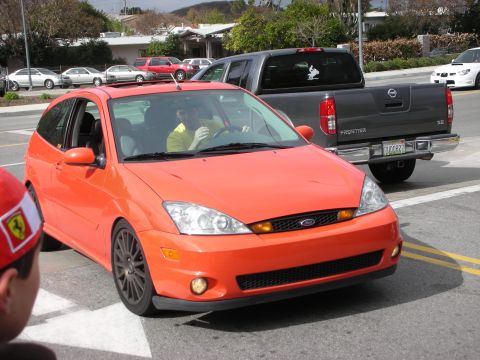 1998 Ford Crown Victoria - orange Ford Focus hatchback with black multi-spoke wheels and fog lights sits in a roadway with onlookers nearby.