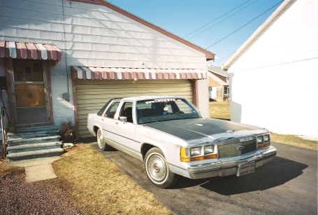 1989 Ford LTD Crown Victoria - A silver 1980s Ford full-size four-door sedan parked in a driveway alongside a house with a striped awning, shown from a 3/4 front angle sh…