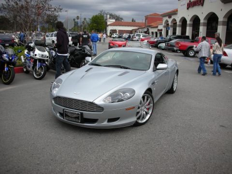 1998 Ford Crown Victoria - silver Aston Martin DB9 coupe with mesh grille parked in a busy shopping-center lot alongside sportbikes.