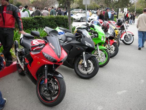 1998 Ford Crown Victoria - row of parked sportbikes in red, green, and black lined up along a hedge with a crowd in the background.