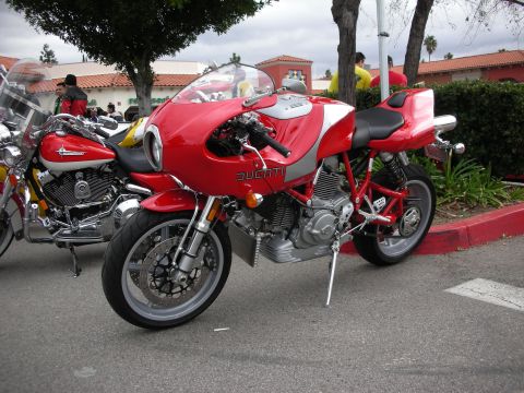 1998 Ford Crown Victoria - red-and-silver Ducati half-faired sportbike parked on its side stand near a curb at an outdoor bike meet.