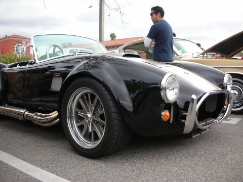 1998 Ford Crown Victoria - black Shelby Cobra-style roadster with polished wheels, side exhaust pipes, and a chrome roll bar at a meet.