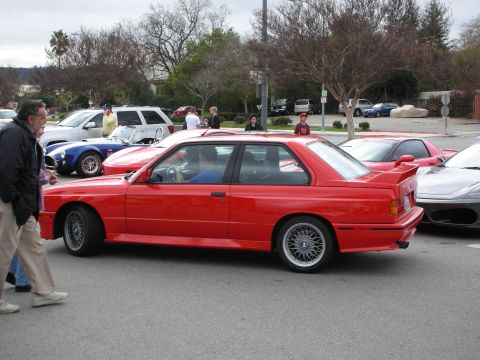1998 Ford Crown Victoria - red BMW E30 M3 coupe shown in side profile in a parking lot as a man walks past at a car show.