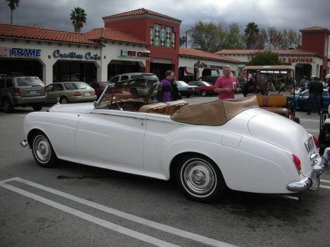1998 Ford Crown Victoria - white classic Rolls-Royce convertible with tan top folded down sits in a strip-mall parking lot at a car show.