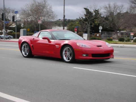 1998 Ford Crown Victoria - red Chevrolet Corvette Z06 coupe in motion on a multi-lane city street under an overcast sky.