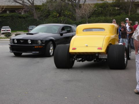1998 Ford Crown Victoria - Rear of a yellow hot rod coupe on wide tires next to a black Ford Mustang on a street at a car gathering.