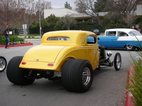 1998 Ford Crown Victoria - Rear three-quarter view of a yellow hot rod coupe with exposed chassis and wide rear tires near a blue classic car.