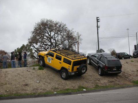1998 Ford Crown Victoria - yellow Hummer H2 with a roof rack sits on a dirt embankment next to a dark SUV as people stand nearby under an overcast sky.