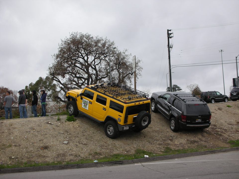Yellow Hummer H2 with roof rack parked on a dirt embankment beside an SUV under gray sky