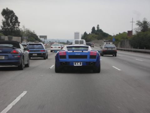 1998 Ford Crown Victoria - blue Lamborghini supercar drives on a multi-lane freeway surrounded by other traffic, seen from behind.