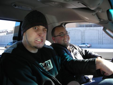 1998 Ford Crown Victoria - Two men sit inside a moving car, one wearing a beanie and making an expression while the other in glasses drives and smiles.