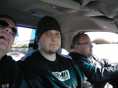 1998 Ford Crown Victoria - Three men sit inside a car, the center one wearing a beanie and dark hoodie, with windows and roadside scenery visible.