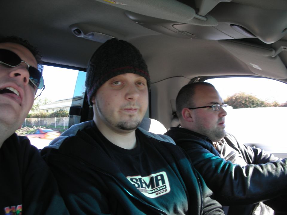 Three men inside a car, center man in a beanie, driver in glasses, with road visible outside