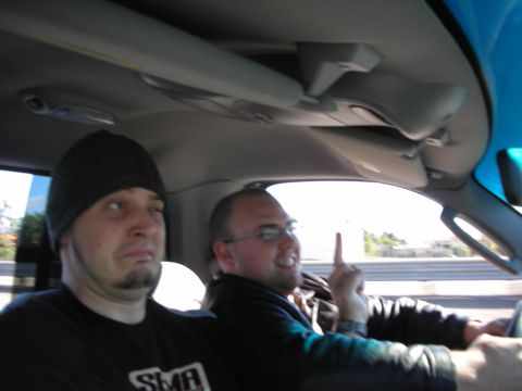 1998 Ford Crown Victoria - Two men ride inside a car on a freeway, one in a beanie and another in glasses raising a finger while driving.