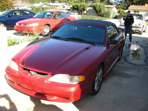 1998 Ford Crown Victoria - red Ford Mustang convertible with a black top and damaged front bumper sits in a driveway as a person stands beside it.