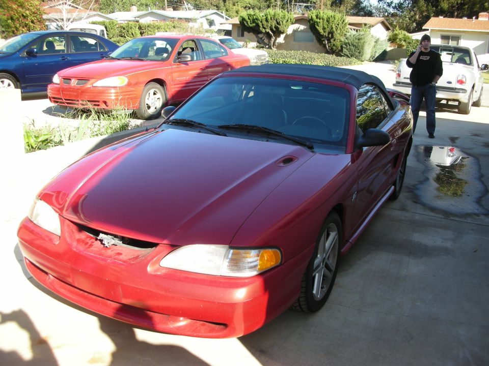 Red Ford Mustang convertible with damaged front bumper parked in a driveway, person nearby
