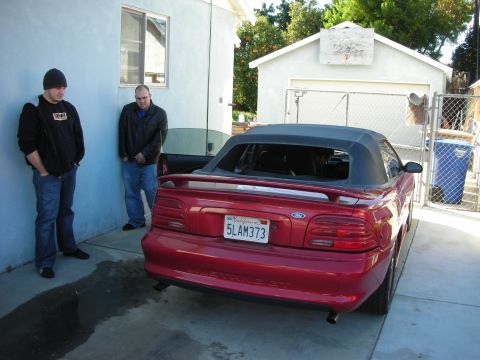 1998 Ford Crown Victoria - red Ford Mustang GT convertible is parked by a stucco wall and chain-link gate with two men nearby; plate reads 5LAM373.