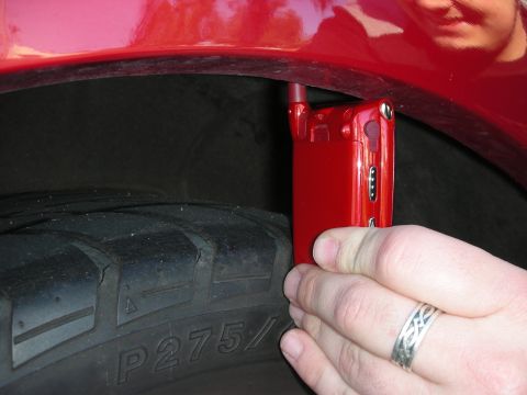 1998 Ford Crown Victoria - hand holds a small red flip phone beside the wheel well of a red car, with a P275-marked tire visible.