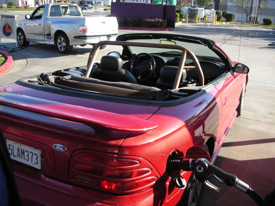 Red Ford Mustang convertible at a gas station fueling, roll bar visible, plate 5LAM373