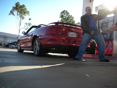 1998 Ford Crown Victoria - low-angle shot of a red Ford Mustang convertible at a gas pump while a man in glasses and a leather jacket fuels it.