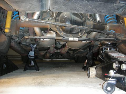 1998 Ford Crown Victoria - car raised on jack stands with a floor jack, exposing the rear axle, dual exhaust and blue coil springs.