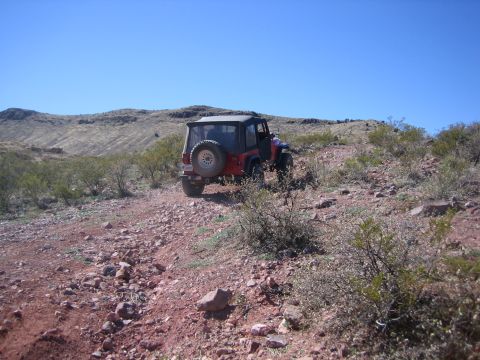 1996 Ford Bronco - A red Jeep Wrangler navigates a rough, rocky terrain on a sunny day with sparse vegetation.
