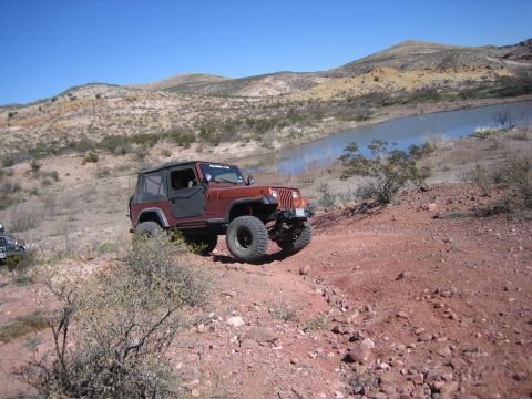 1996 Ford Bronco - A brown Jeep Wrangler navigates a rocky, off-road trail near a body of water under a clear blue sky.