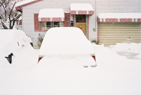 1989 Ford LTD Crown Victoria - Two cars buried under deep snow in a driveway after a heavy snowfall, parked in front of a house with pink-and-white striped window awnings…