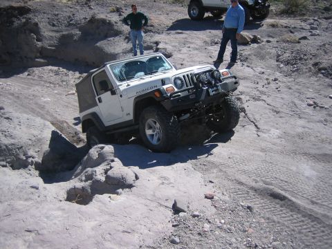 1996 Ford Bronco - A white Jeep Wrangler Rubicon is shown off-roading on a rocky, dusty trail with two onlookers.