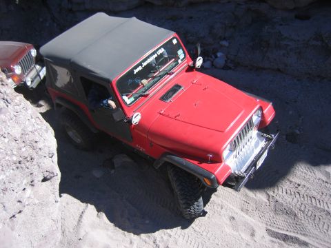 1996 Ford Bronco - A red Jeep Wrangler navigates a rocky, sandy canyon with another vehicle visible behind it.