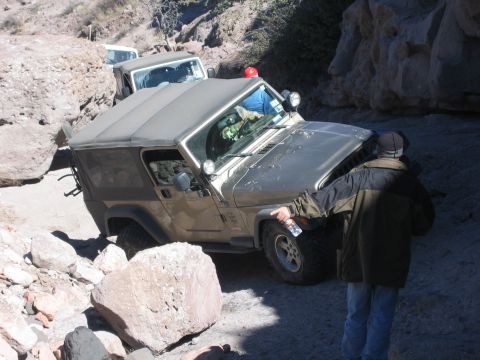 1996 Ford Bronco - Gold Jeep Wrangler is being guided by a person on a difficult rocky trail, with a white Jeep following behind.