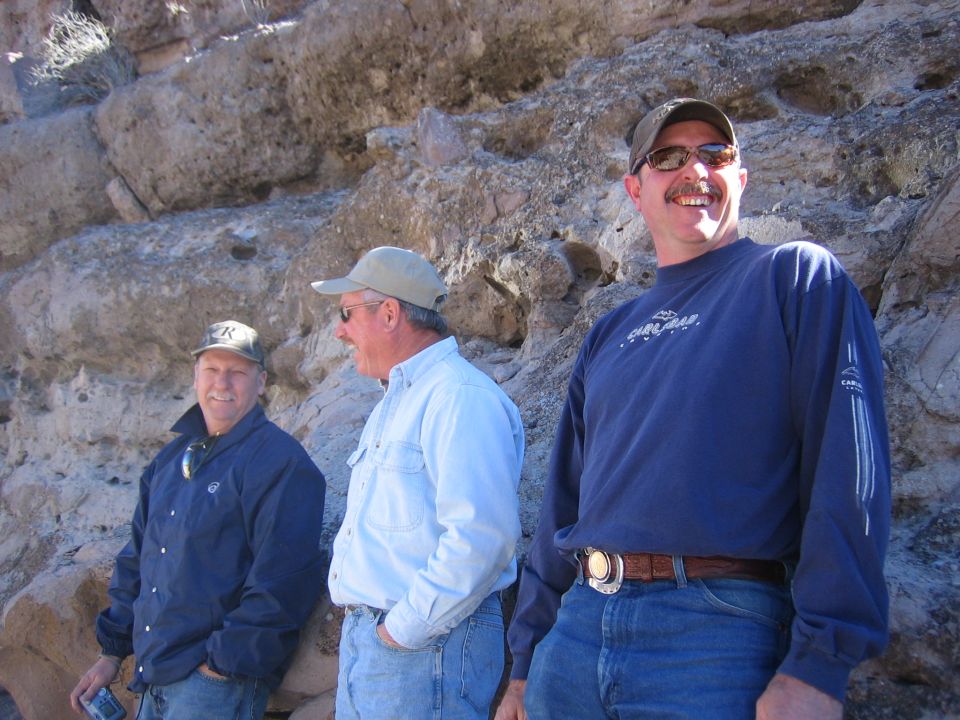 Three men stand in front of a rocky cliff face. The man on the right wears a blue long-sleeved shirt and jeans. The man in the middle wears a light blue button-up shirt and jeans. The man on the left wears a dark blue jacket and jeans